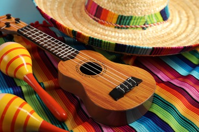 Photo of Mexican sombrero hat, maracas and ukulele on color poncho, closeup