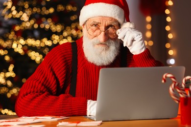 Photo of Santa Claus using laptop at his workplace in room decorated for Christmas