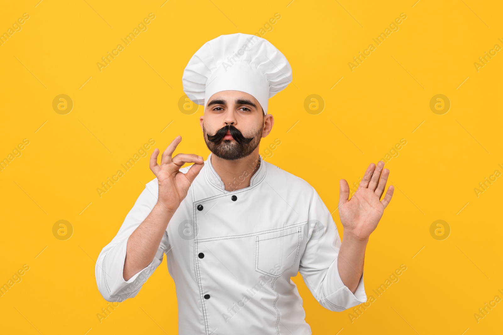 Photo of Professional chef with funny artificial moustache showing perfect sign on yellow background
