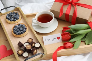 Photo of Tasty breakfast served in bed. Delicious desserts, tea, flowers, gift box and card with phrase I Love You on tray