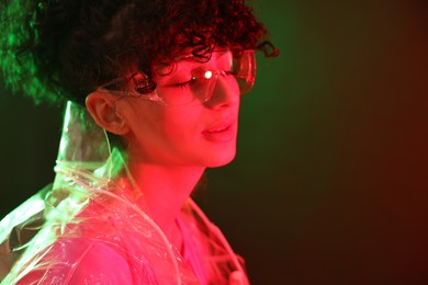 Photo of Beautiful young woman in transparent coat and sunglasses posing on color background in neon lights. Space for text