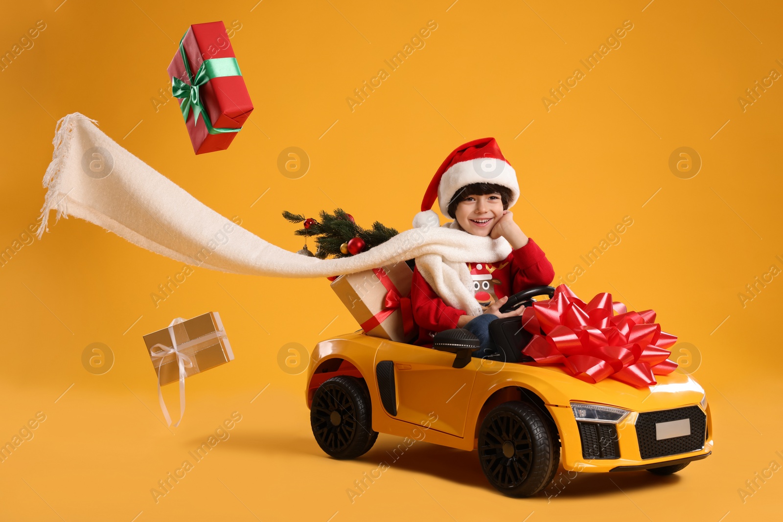 Photo of Cute little boy in Santa hat with Christmas tree and gift boxes driving children's electric toy car on yellow background