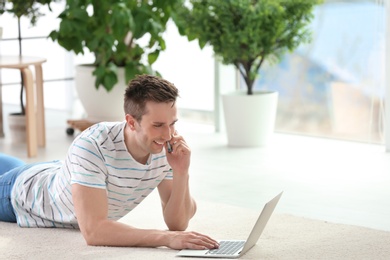 Photo of Handsome young man with mobile phone and laptop on cozy carpet at home