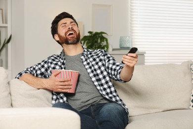 Laughing man watching TV with popcorn on sofa indoors