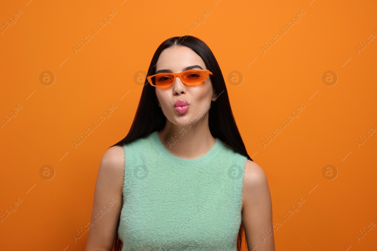 Photo of Beautiful young woman in stylish sunglasses blowing kiss on orange background