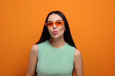 Beautiful young woman in stylish sunglasses blowing kiss on orange background