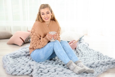Photo of Attractive young woman in cozy warm sweater with cup of hot drink sitting on floor at home