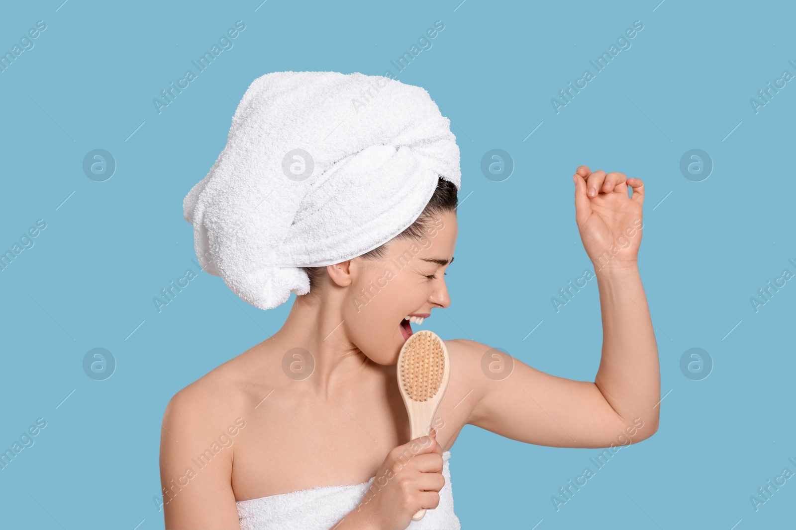 Photo of Emotional woman in towels singing into brush on light blue background. Spa treatment