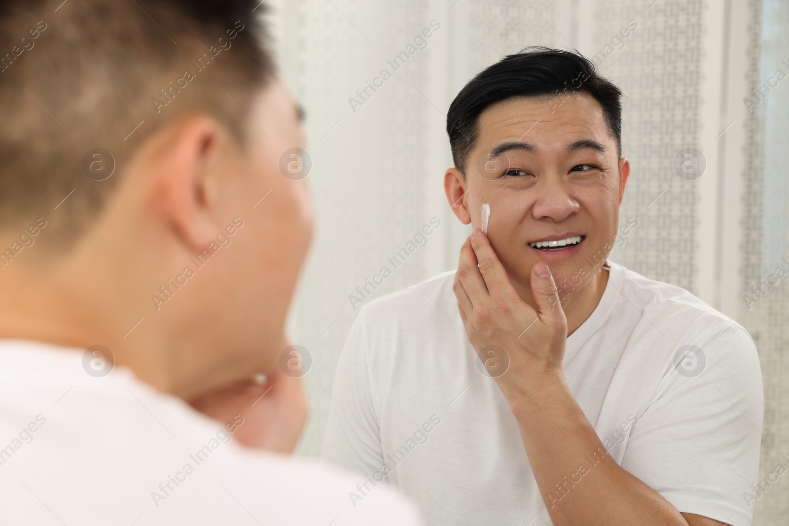 Photo of Handsome man applying cream onto his face near mirror indoors