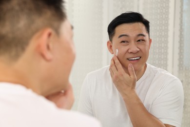 Handsome man applying cream onto his face near mirror indoors