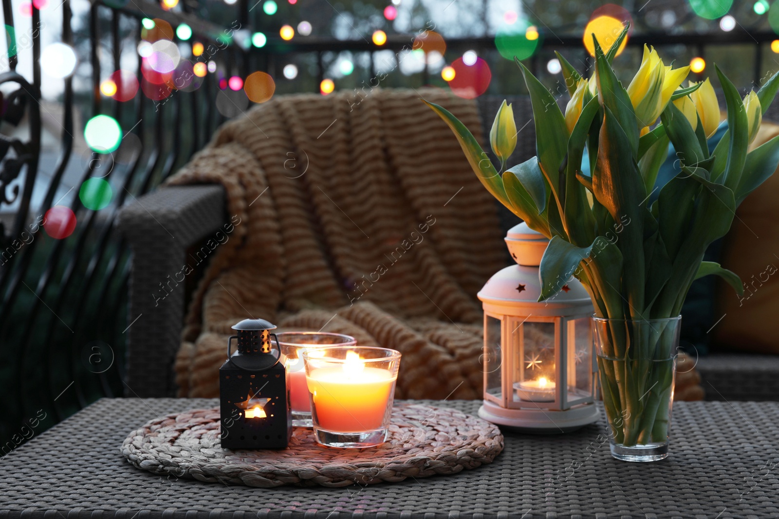 Photo of Soft blanket, burning candles and yellow tulips on rattan garden furniture in evening