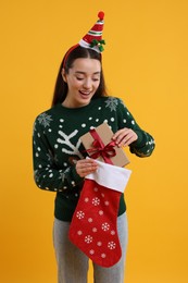 Happy young woman in Christmas sweater taking gift from stocking on orange background