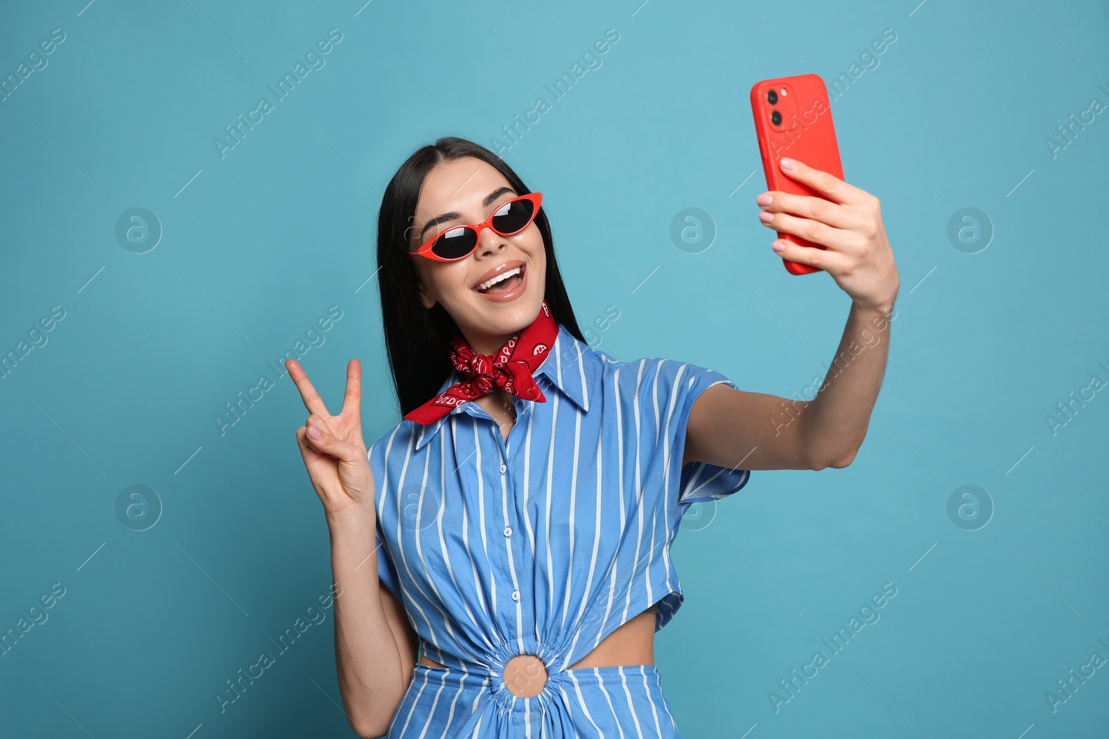 Photo of Fashionable young woman in stylish outfit with bandana taking selfie on light blue background