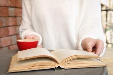 Woman with cup of coffee reading book at table outdoors, closeup