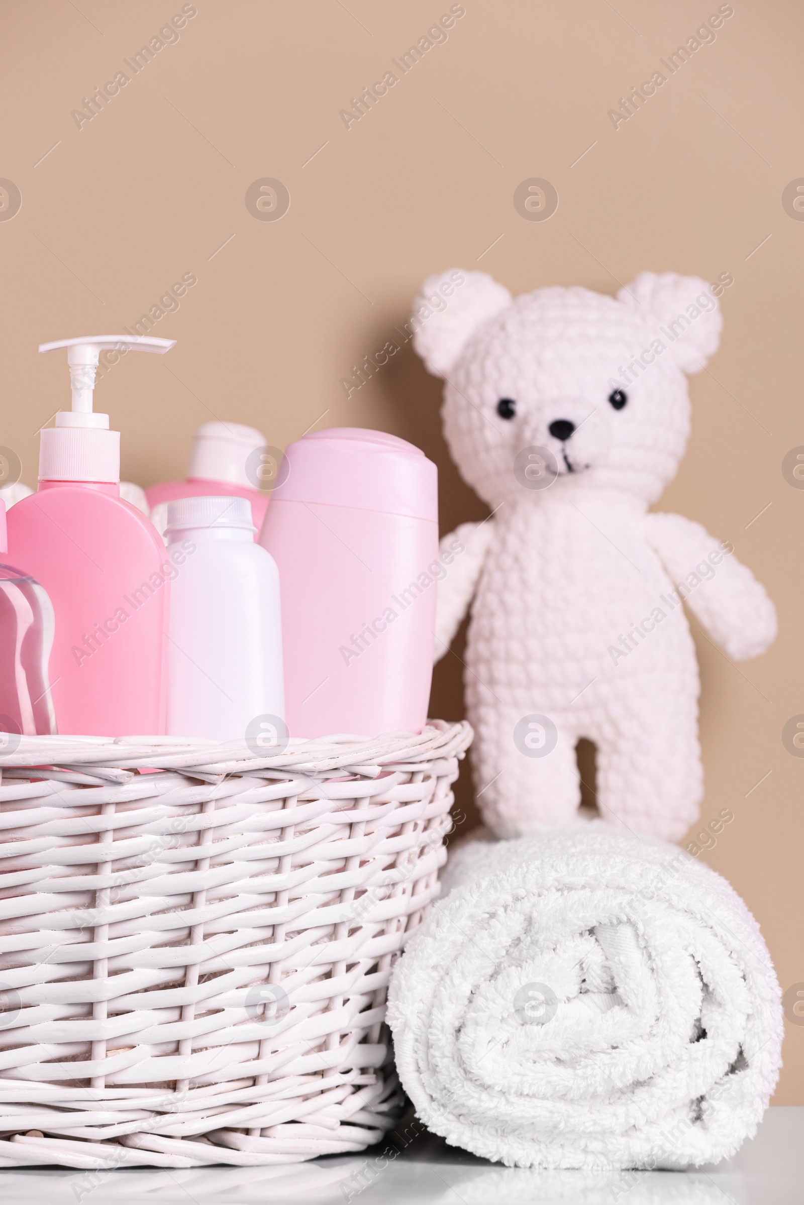 Photo of Wicker basket with baby cosmetic products, bath accessories and knitted toy bear on white table against beige background