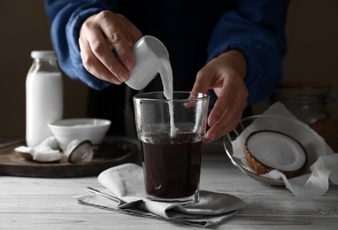 Photo of Woman pouring coconut milk into glass of coffee at white wooden table, closeup