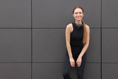 Photo of Beautiful woman in stylish gym clothes with headphones near dark grey wall on street, space for text