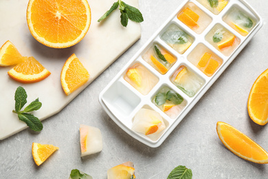 Photo of Ice cubes in tray with orange slices and mint on light grey table, flat lay
