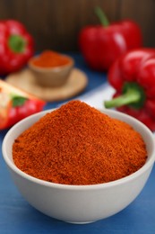 Photo of Bowl with aromatic paprika powder and fresh bell peppers on blue wooden table