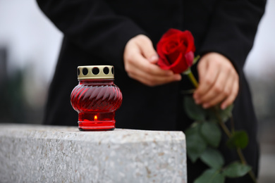 Woman holding red rose near grey granite tombstone with candle outdoors, closeup. Funeral ceremony