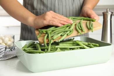 Photo of Woman pouring green beans into baking dish at white table, closeup