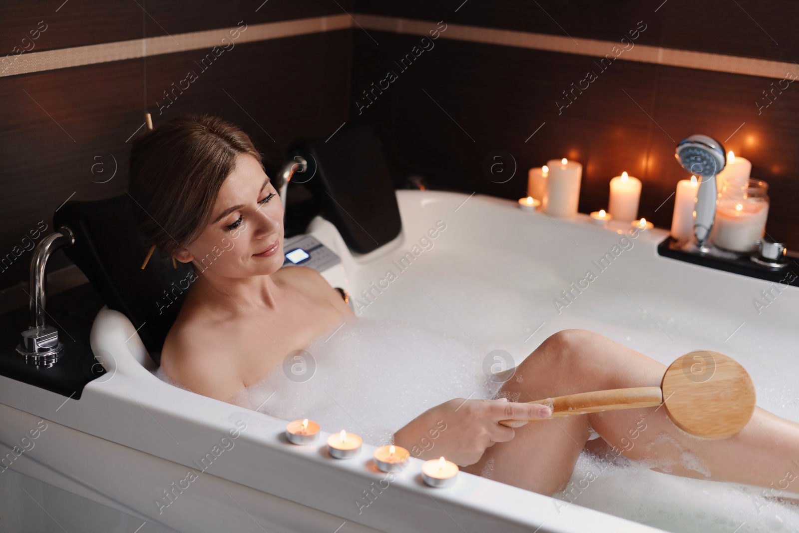 Photo of Beautiful woman rubbing leg with brush while taking bubble bath indoors. Romantic atmosphere