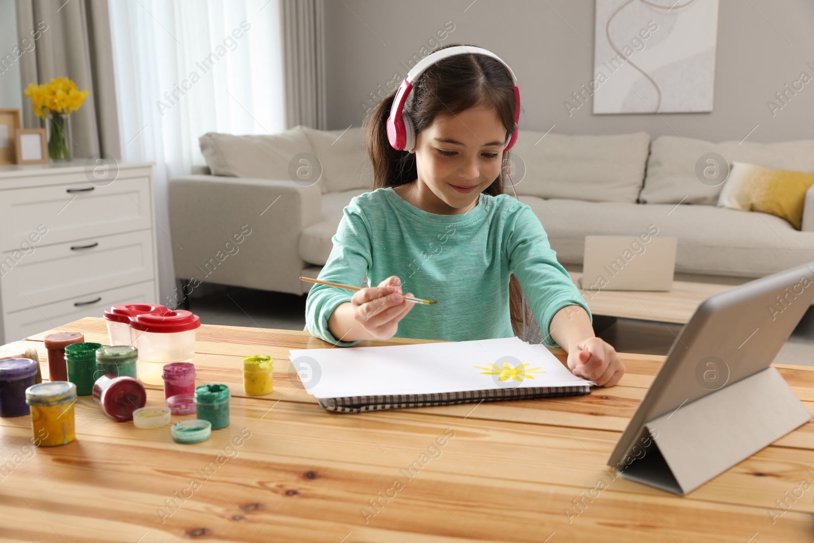 Photo of Little girl with headphones drawing on paper at online lesson indoors. Distance learning