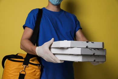 Courier in protective gloves holding pizza boxes on yellow background, closeup. Food delivery service during coronavirus quarantine