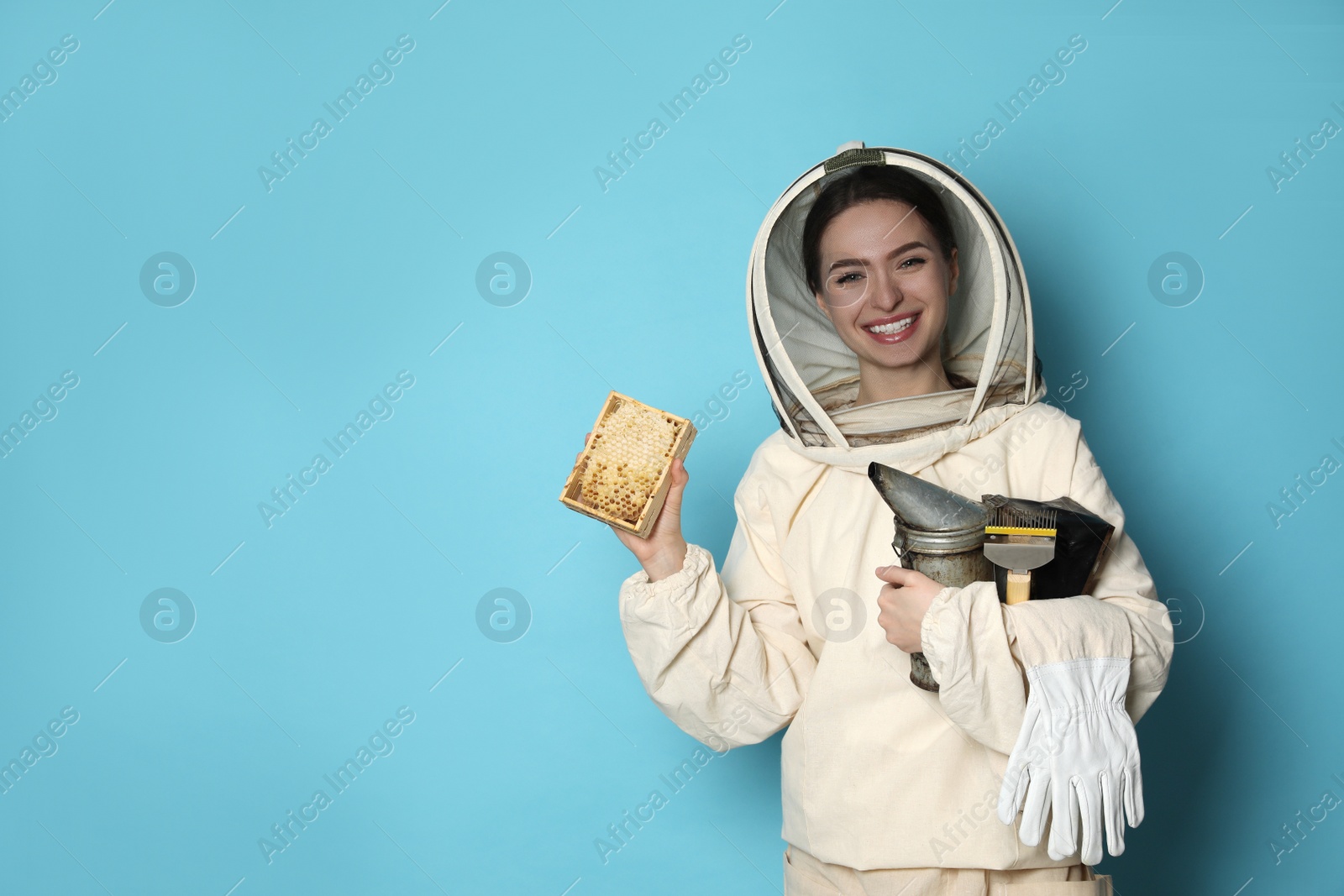Photo of Beekeeper in uniform holding smokepot and hive frame with honeycomb on light blue background. Space for text