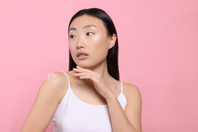 Beautiful young Asian woman with smear of body cream on shoulder against pink background