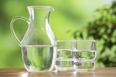 Jug and glasses with clear water on wooden table against blurred green background, closeup