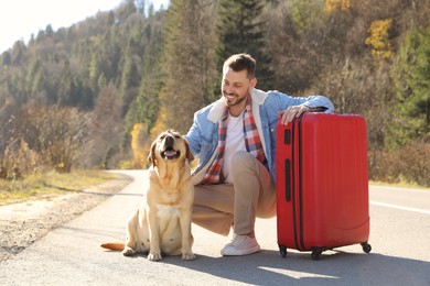 Happy man with adorable dog and red suitcase on road. Traveling with pet