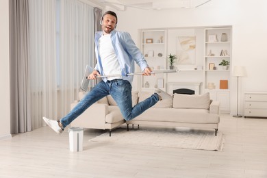 Photo of Enjoying cleaning. Man in headphones jumping with mop at home