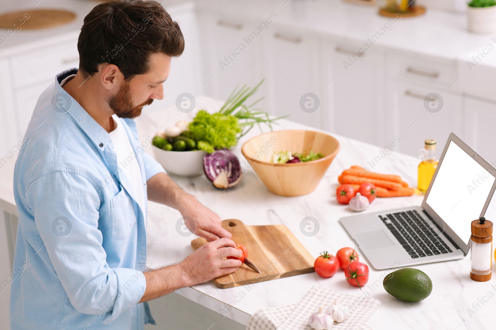 Photo of Man making dinner while watching online cooking course via laptop in kitchen