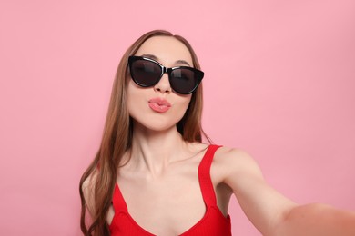 Photo of Beautiful young woman with sunglasses blowing kiss while taking selfie on pink background