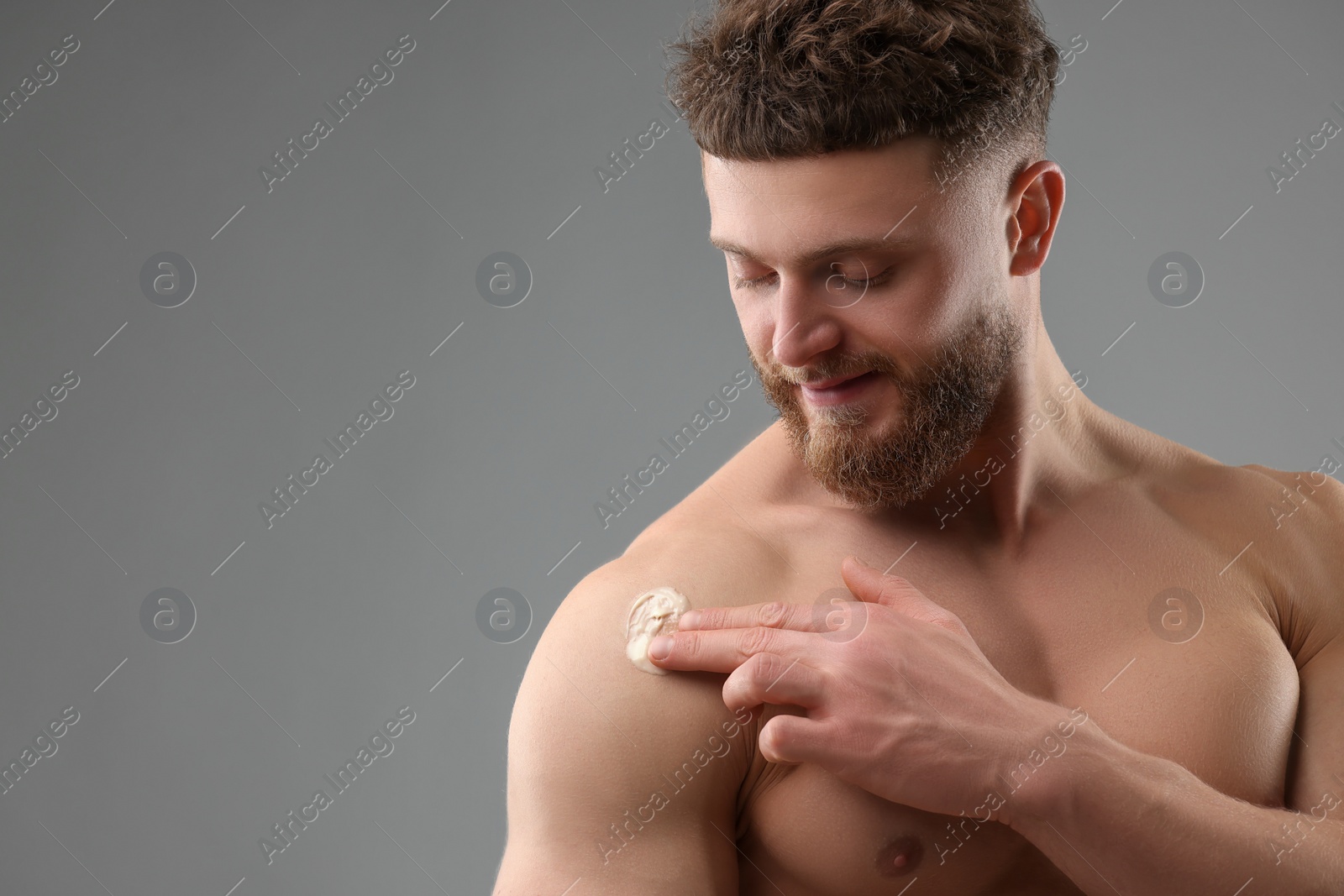 Photo of Handsome man applying moisturizing cream onto his shoulder on grey background, space for text