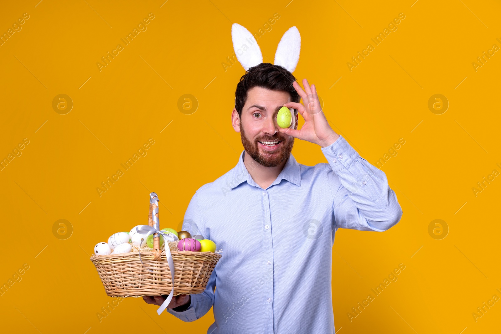 Photo of Portrait of happy man in cute bunny ears headband holding wicker basket and covering eye with Easter egg on orange background