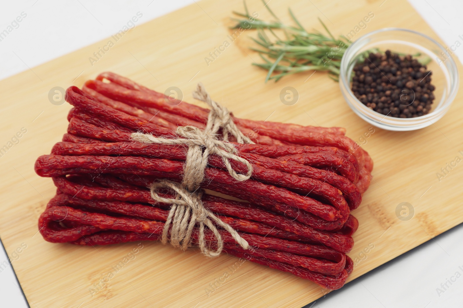 Photo of Bundles of delicious kabanosy with peppercorn and rosemary on white wooden table, closeup