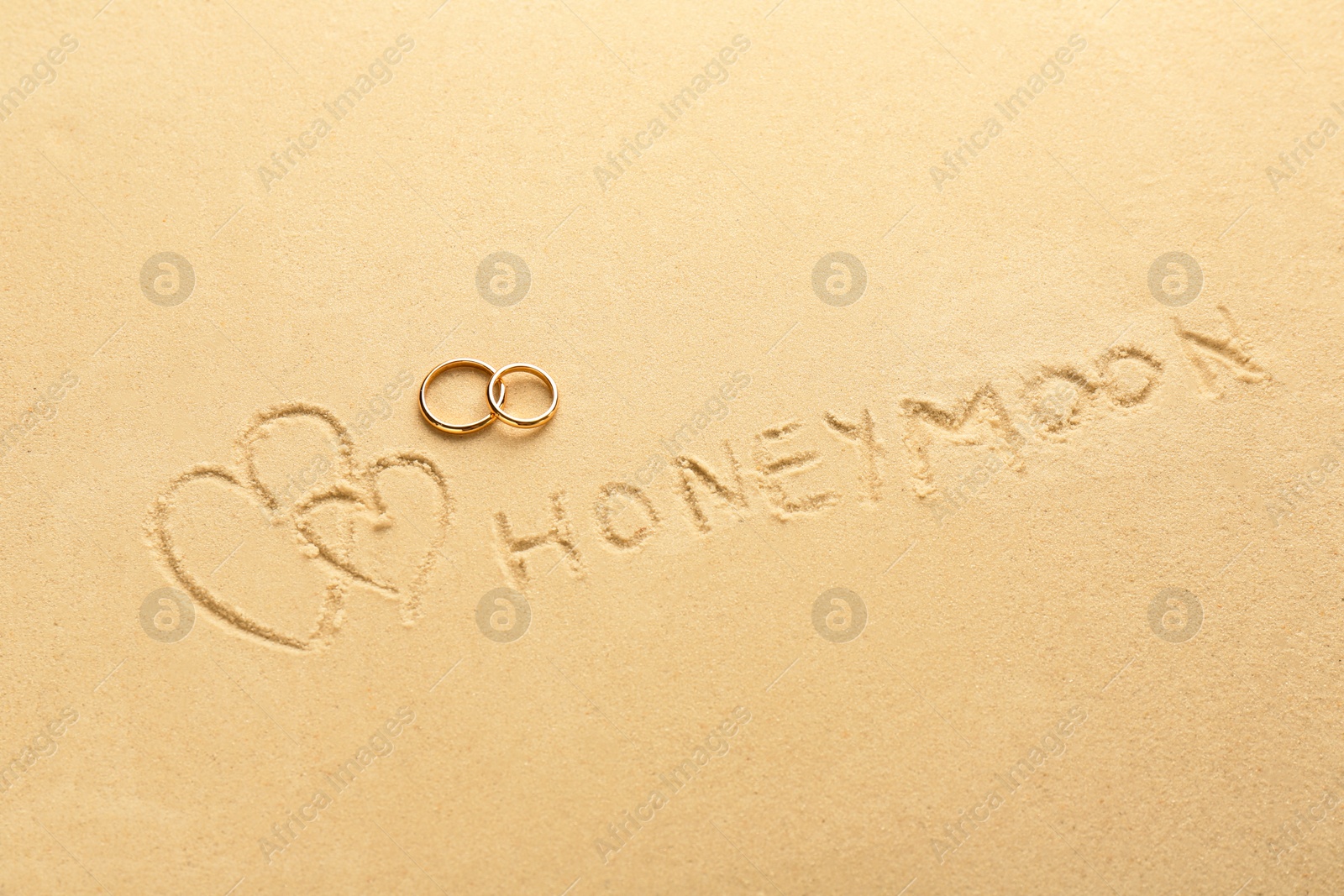 Photo of Word Honeymoon written on sand, hearts two golden rings, top view