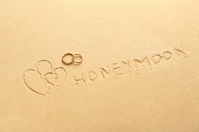 Word Honeymoon written on sand, hearts two golden rings, top view