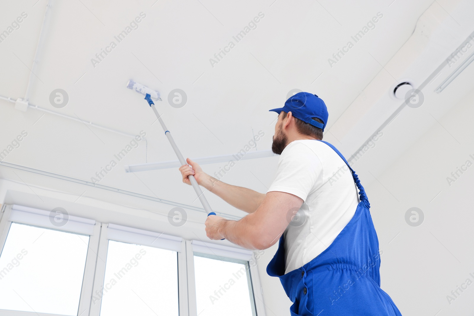 Photo of Worker in uniform painting ceiling with roller on stepladder indoors, low angle view