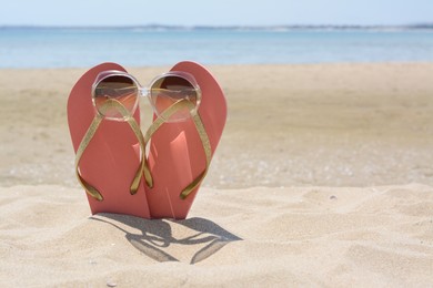 Stylish pink flip flops with sunglasses in sand near sea on sunny day, space for text