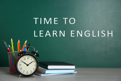 Alarm clock and stationery on table near green chalkboard with text Time to Learn English