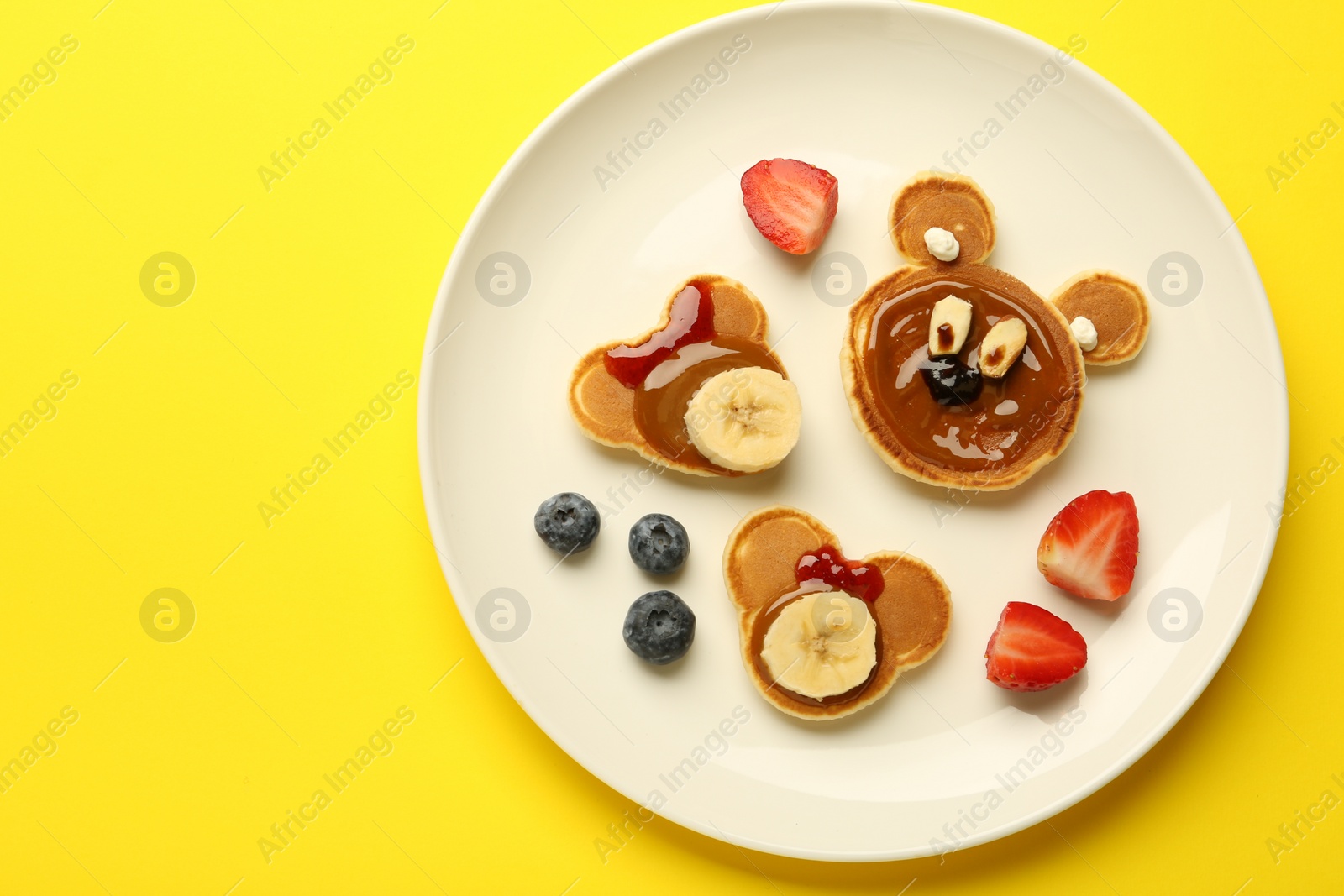 Photo of Creative serving for kids. Plate with cute bears made of pancakes, berries, banana and chocolate paste on yellow background, top view. Space for text