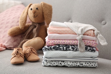 Toy bunny, stack of stylish child clothes and shoes on sofa