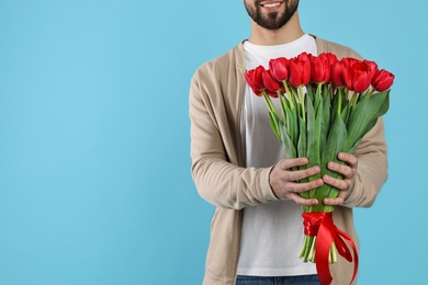 Photo of Happy man holding red tulip bouquet on light blue background, closeup view with space for text. 8th of March celebration