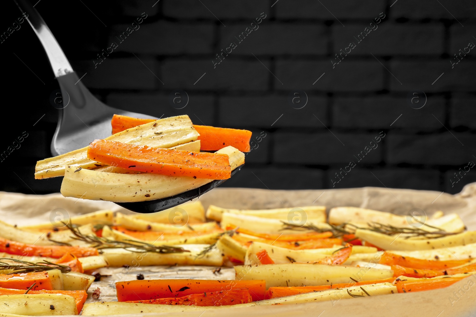 Photo of Taking baked parsnip and carrot with spatula from tray against brick wall, closeup