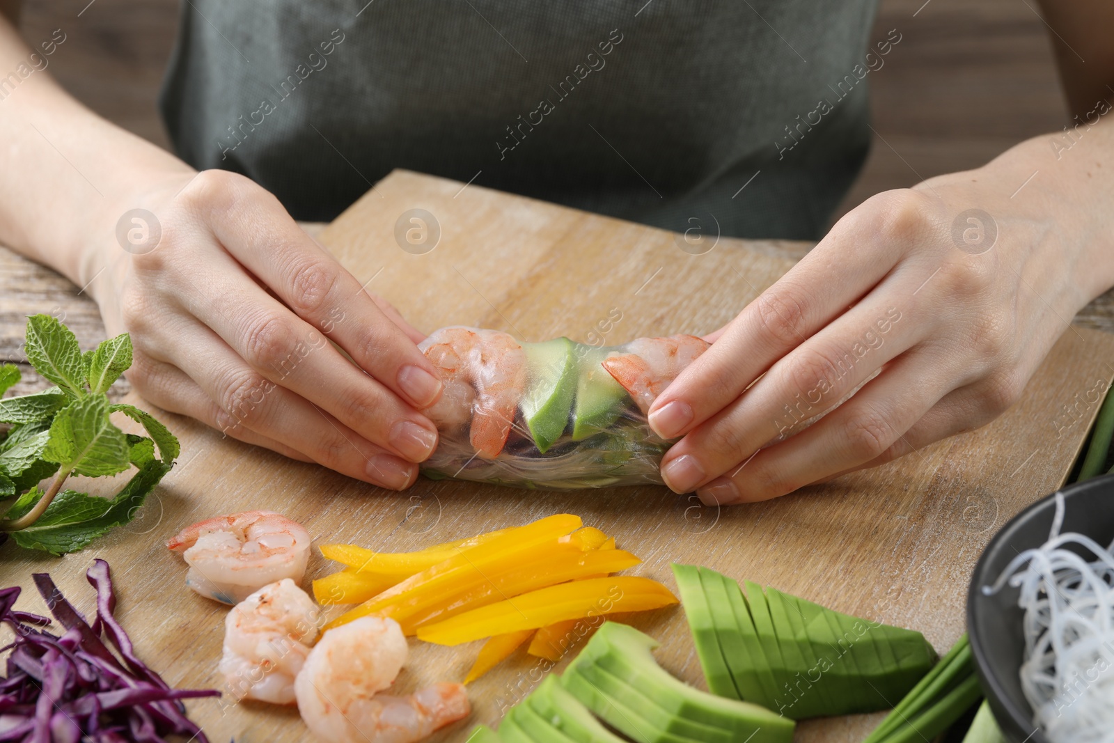 Photo of Woman wrapping spring roll at table with products, closeup