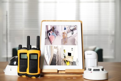 Tablet with view from CCTV cameras, walkie talkies, smoke and movement detectors on wooden table indoors. Home security system