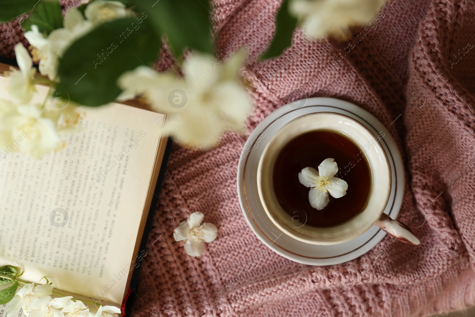 Photo of Cup of aromatic tea with beautiful jasmine flower and open book on pink fabric, flat lay